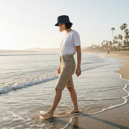 Woman wearing a Fog & Foundry dark grey cotton twill bucket hat while walking barefoot along a sunny beach.