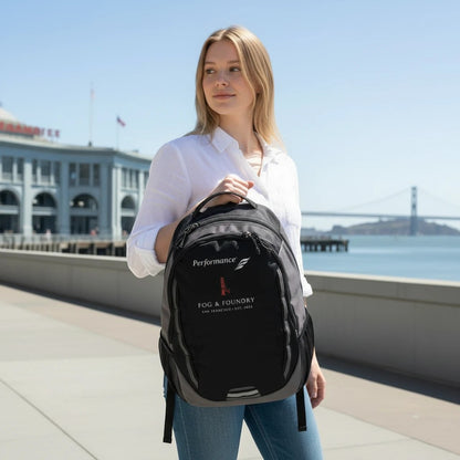Woman carrying a black Fog & Foundry Ridge backpack with embroidered hummingbird logo on a sunny San Francisco pier.