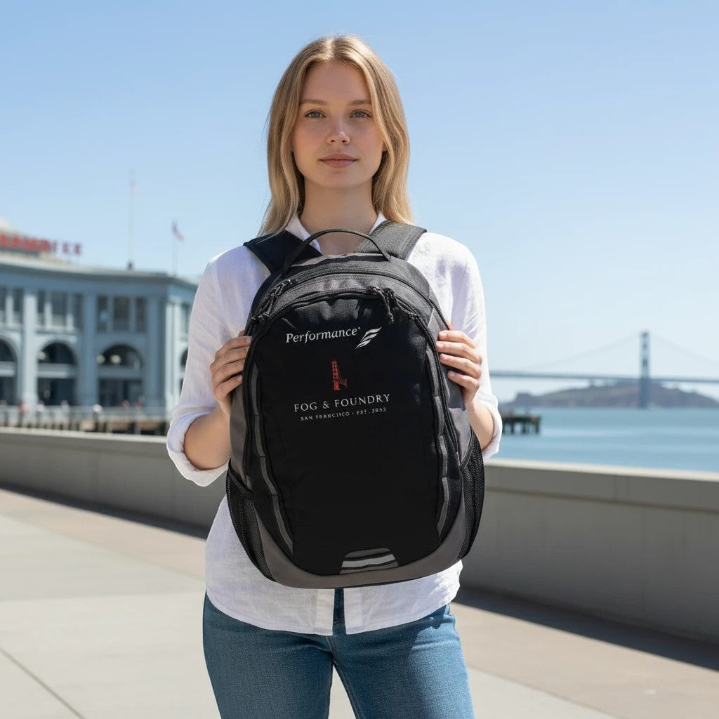 Woman wearing a black Fog & Foundry Ridge backpack with embroidered hummingbird detail outdoors by the bay.