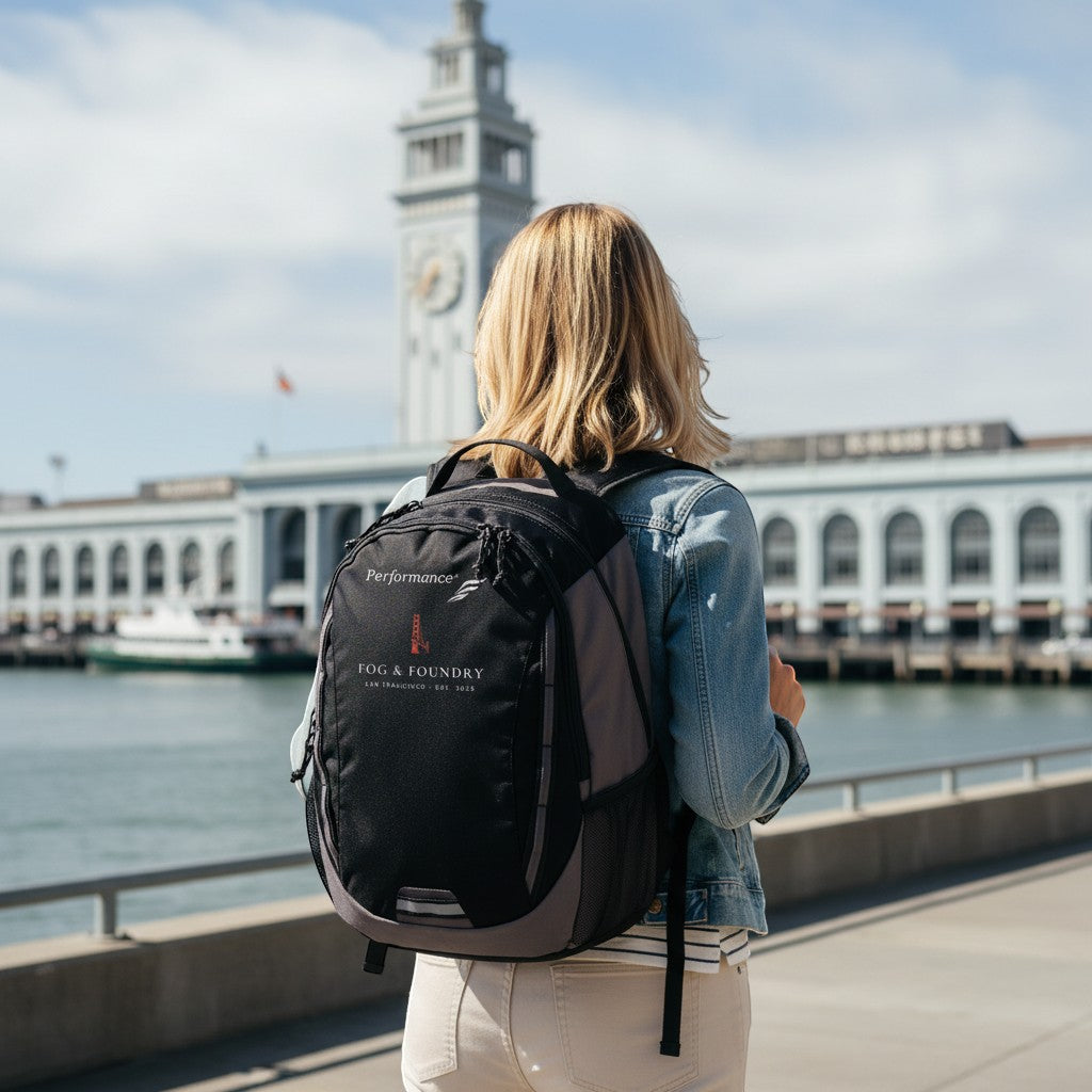 Woman wearing a black Fog & Foundry Ridge backpack with embroidered hummingbird logo near the San Francisco waterfront.
