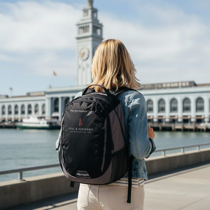 Woman wearing a black Fog & Foundry Ridge backpack with embroidered hummingbird logo near the San Francisco waterfront.