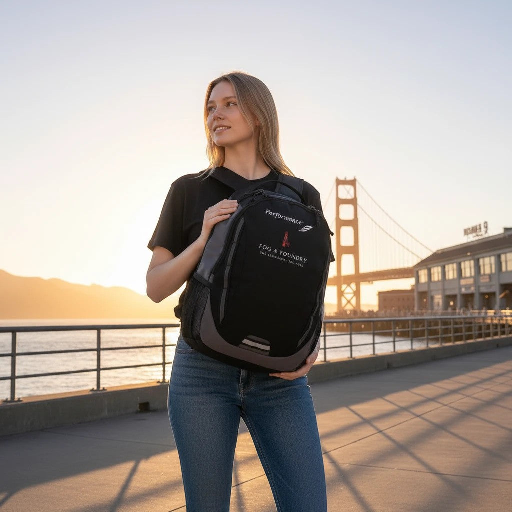 Woman holding a black Fog & Foundry Ridge backpack with embroidered logo, standing before the Golden Gate Bridge.
