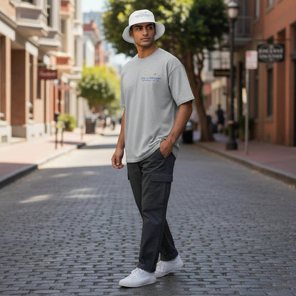 A man wearing a Fog & Foundry white cotton twill bucket hat and gray t-shirt on a cobblestone city street.