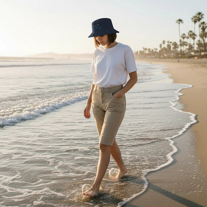 A woman wearing a dark gray Fog & Foundry cotton twill bucket hat while walking barefoot along a sunny beach.