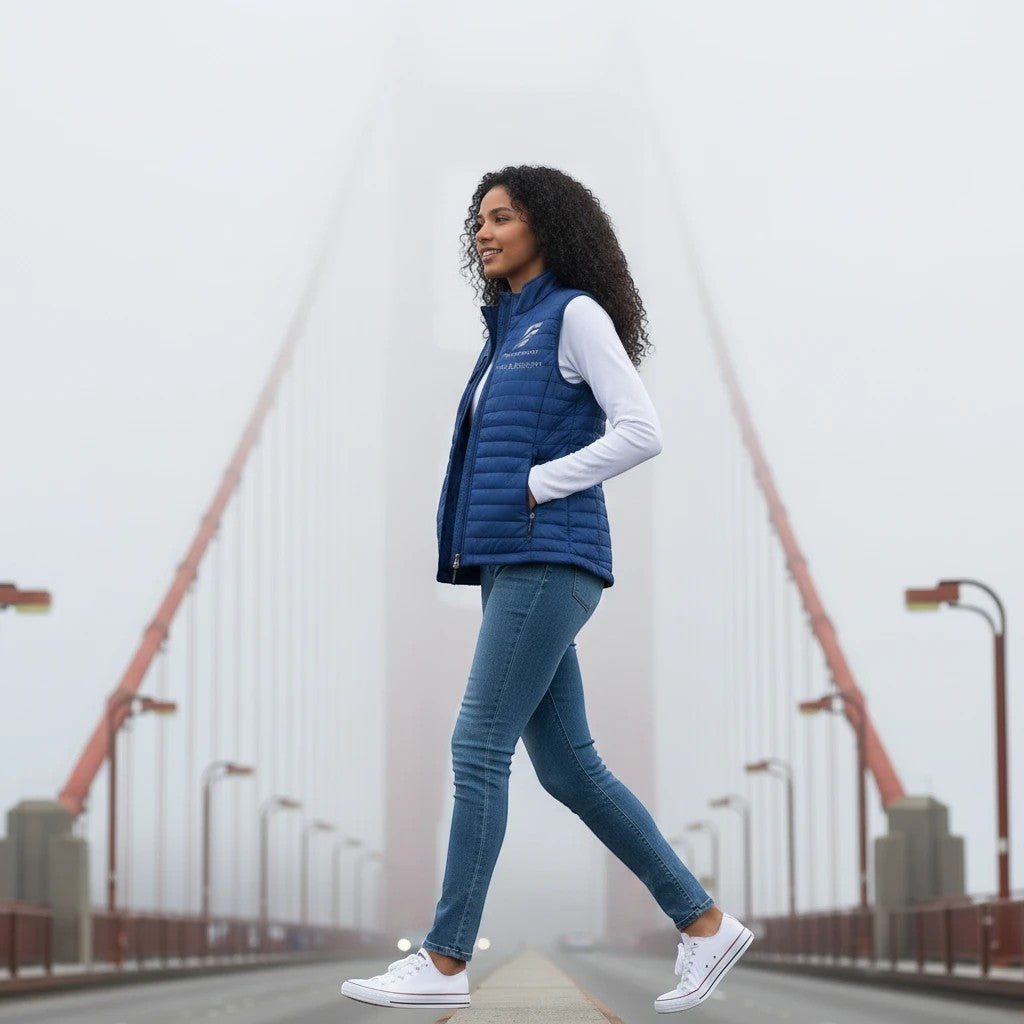 A woman wearing a blue Fog & Foundry quilted Marina Vest with a hummingbird logo, walking on a foggy bridge.