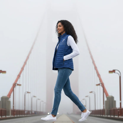 A woman wearing a blue Fog & Foundry quilted Marina Vest with a hummingbird logo, walking on a foggy bridge.