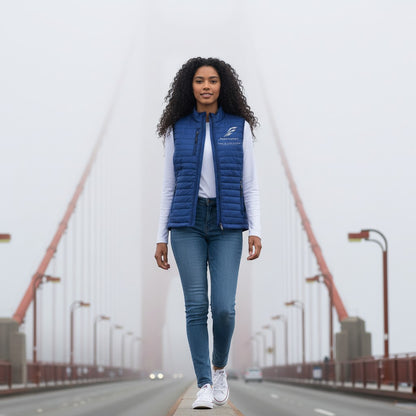 A woman wearing a blue Fog & Foundry quilted Marina Vest with a hummingbird logo, walking on a foggy bridge.
