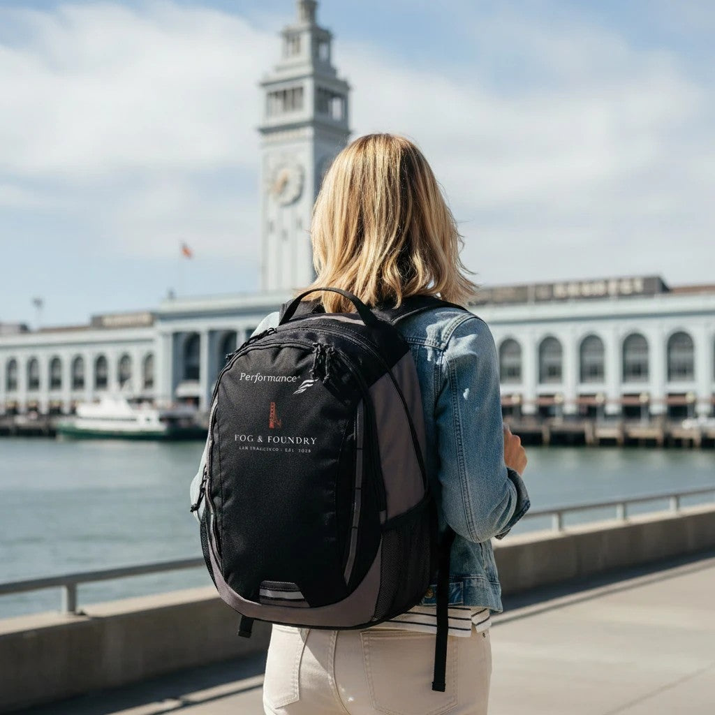 Woman wearing a black Fog & Foundry Ridge backpack with embroidered hummingbird logo at San Francisco Ferry Building