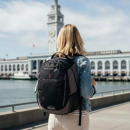Woman wearing a black Fog & Foundry Ridge backpack with embroidered hummingbird logo at San Francisco Ferry Building