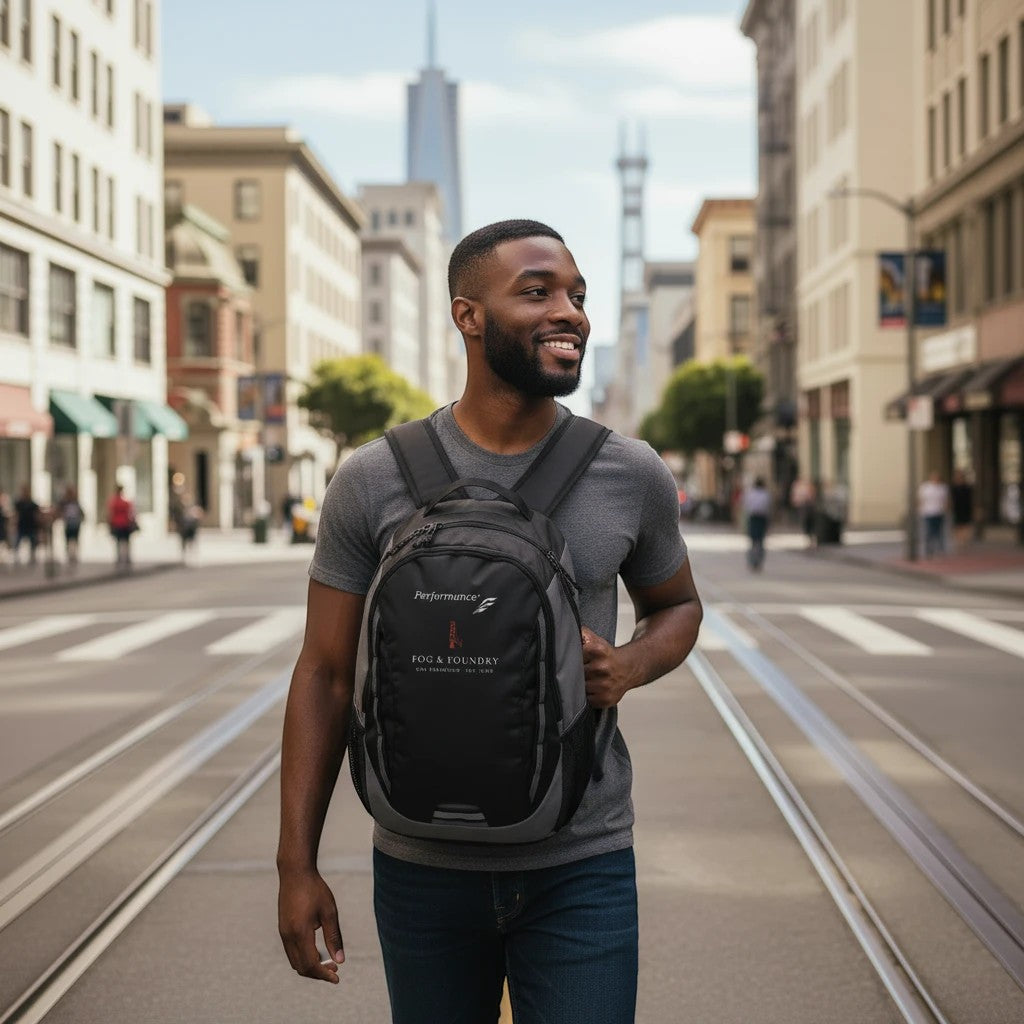 Man wearing a Fog & Foundry black Ridge backpack while walking on a city street with urban buildings in the background