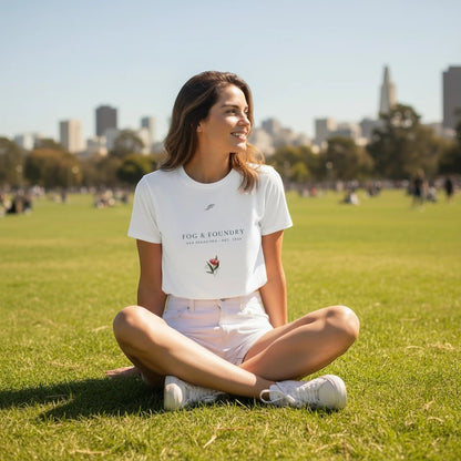 Woman wearing a white Fog & Foundry crop tee with a hand-drawn bloom graphic, sitting on a sunny city park lawn.