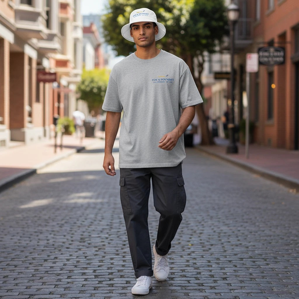 A man wearing a Fog & Foundry white cotton bucket hat and gray t-shirt walking down a city street.