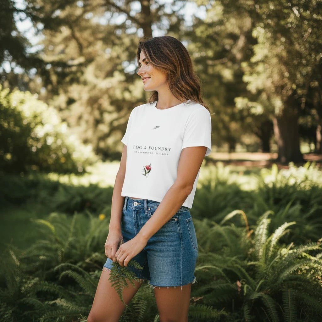 Woman wearing a white Fog & Foundry crop tee with a hand-drawn bloom graphic in a lush, sunlit garden setting.